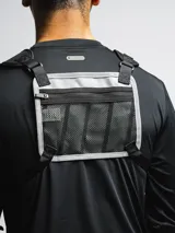 Bolso de pecho táctico color gris claro con detalles en negro, con correas ajustables y hombreras de neopreno. Cuenta con tres compartimentos: uno trasero de red y dos frontales con cierre.