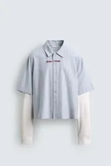 Camisa de corte relajado con diseño de rayas verticales en tonos azul y blanco. Presenta cuello solapa, cierre frontal de botones y un detalle de texto bordado en color rojo en el pecho. Cuenta con un diseño de manga larga con efecto doble, combinando la manga corta de la camisa con una manga larga de tejido tipo waffle en color crudo.