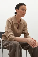 Sweater de punto color beige con cuello polo, escote en V y mangas largas. Presenta un diseño de estructuras trenzadas en el tejido.