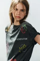 Camiseta oversize de algodón con efecto lavado, cuello redondo y manga corta. Presenta múltiples aplicaciones de brillos y strass con motivos coloridos de Smiley, incluyendo estrellas, corazones y caras sonrientes.
