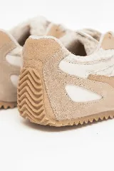 Championes deportivos en tonos beige y ocre, confeccionados con combinaciones de materiales texturados y forro interior de corderito sintético. Presentan suela dentada de goma que se extiende hacia el talón.