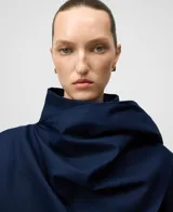 Cazadora corta de gabardina azul marino con cuello chimenea y cierre frontal cruzado con botones. Presenta un diseño asimétrico con una pieza de tejido integrada en la manga para llevar a modo de chal.