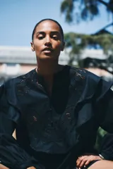 Vestido corto negro, tipo túnica, confeccionado en mezcla de lino y algodón. Presenta inserciones de encaje calado en el canesú y volados superpuestos en el frente y hombros. Tiene escote redondo, cierre frontal con botones forrados y mangas largas con puños finos abotonados.