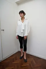 Camisa de manga larga confeccionada en satén de color blanco, con superficie lisa, leve brillo y caída suave. Presenta cuello clásico y cartera frontal.
