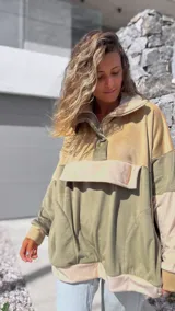 Buzo tipo canguro de diseño color block en tonos verde oliva y beige, confeccionado en tejido de textura aterciopelada. Presenta cuello alto con cierre frontal parcial, capucha y un bolsillo frontal grande con solapa.