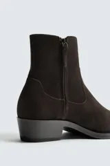 Bota estilo cowboy de caña media, confeccionada en piel con acabado de gamuza en color marrón oscuro. Presenta horma en punta, cierre lateral y suela con tacón cuadrado bajo.