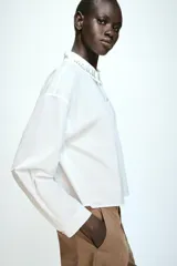 Camisa corta de popelín en color blanco, con cuello solapa decorado con abalorios joya, manga larga y cierre frontal con botones.