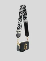 Bolso bandolera tipo cámara (camera bag) Marc Jacobs The Snapshot, color negro con detalles en blanco y herrajes dorados. Presenta el logo 'J' en metal dorado en el frente y una correa ancha de tela con el logo de la marca en blanco y negro.