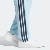 Pantalón deportivo Adidas de corte clásico, color celeste con las icónicas tres rayas laterales en color azul oscuro. Presenta cintura elástica con cordón de ajuste, bolsillos laterales y diseño de pierna recta con puños abiertos.