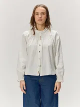 Camisa de algodón color crudo, con cuello alto fruncido, cierre frontal abotonado y mangas largas con puños elásticos fruncidos.