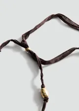 Collar con cinta marrón ajustable en la parte posterior, abalorios dorados texturizados a lo largo de la tira y un dije circular central dorado con diseño de espiral en relieve y pequeños cristales incrustados.