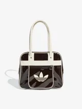 Bolso tipo shopper de Adidas, confeccionado en material sintético con acabado de charol brillante en color marrón oscuro y detalles en blanco. Presenta el logo del Trifolio estampado en el frente, bolsillo exterior con cierre y asas largas para llevar al hombro.