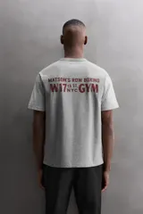 Camiseta relaxed fit de algodón, color gris melange con mangas en un tono de gris más claro. Tiene cuello redondo, manga corta, bordado frontal y texto estampado en la espalda a contraste. Incluye un parche con logo en el bajo.