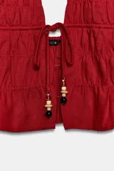 Top halter corto de color rojo, confeccionado con lino y tejido fruncido. Posee escote en V pronunciado, espalda descubierta, tira elástica trasera y cierre frontal con lazo decorado con abalorios colgantes.