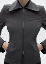 Chaqueta entallada de color gris con diseño de rayas verticales finas. Presenta cuello camisero, cierre frontal de cremallera doble y bolsillo de ribete en el pecho.
