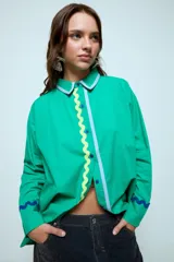 Camisa azul de corte oversize, con cierre frontal de botones y detalles de cintas onduladas en colores contrastantes a lo largo de la botonadura y en los puños. Presenta cuello clásico y mangas largas.