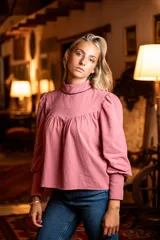 Blusa rosa de algodón con cuello alto y mangas largas abullonadas. Presenta detalles de cinta negra en el cuello y los puños, hombros con diseño princesa y cierre de lágrima con botón forrado en la parte posterior del cuello.