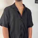 Camisa de tejido calado con textura, color blanco, de corte oversize, manga corta y cuello solapa con cierre frontal de botones.