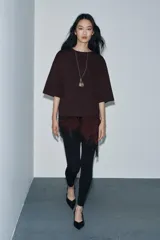 Camiseta de algodón de corte regular, color marrón oscuro o negro, con cuello redondo y manga corta. Presenta un bajo asimétrico con aplicación de encaje negro a tono.