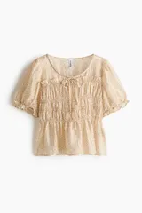 Blusa de lyocell con estampado floreado pequeño sobre base beige, escote redondo con tiras decorativas para atar, busto fruncido, mangas cortas abullonadas con elástico y ribete de volado, y bajo levemente acampanado.