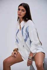 Camisa blanca de manga larga con cuello clásico y botones frontales. Presenta un diseño de ondas en tonos azules con lentejuelas y pequeños apliques metálicos en el frente.