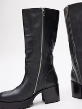 Bota bucanera de cuero vacuno wax color negro, con punta cuadrada, suela de goma con plataforma dentada y taco de madera. Presenta detalle de doble hebilla metálica en la parte superior de la caña.