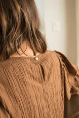Blusa de tela texturizada color café, con cuello redondo y mangas al codo que presentan tres volados superpuestos y puños elásticos. Cuenta con cierre de botón en la parte posterior.