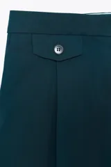 Pantalón de vestir marrón oscuro, tiro alto, con pernera ancha y pinzas frontales. Presenta un bolsillo con tapeta en la parte superior delantera.