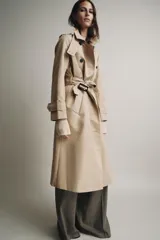 Trench color beige con cuello solapa y manga larga ajustable con trabillas. Bolsillos en delantero. Cinturón en mismo tejido. Detalle de pespuntes marcados a tono. Cierre frontal cruzado con botones. Tejido repelente al agua.