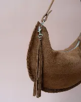 Bolso de hombro tipo hobo confeccionado en gamuza suave color marrón oscuro. Presenta un diseño de media luna con bordes trenzados, cierre superior con tiradores de cuero anudado y una correa ajustable y desmontable con herrajes metálicos.