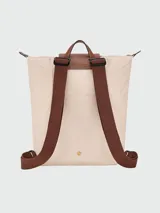 Mochila de diseño minimalista en color beige, con solapa y asa superior en cuero marrón texturizado. Cuenta con correas ajustables en color marrón y cierre principal.