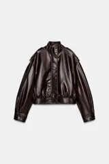 Cazadora corta de efecto piel color marrón oscuro, con cuello subido y cierre frontal oculto con cremallera y botones a presión. Presenta mangas largas con puños y pliegues, trabillas en los hombros, bolsillos laterales y bajo abullonado. El tejido tiene un efecto lavado.