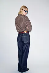 Camisa de manga larga con cuello subido, cierre frontal con botones y detalle de volantes en el pecho. Presenta un diseño a rayas verticales y puños elásticos.