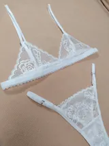 Conjunto de lencería de dos piezas confeccionado en encaje blanco con diseño floral. Incluye un sostén triangular con tirantes regulables y una bombacha colaless ajustable con tiras laterales.