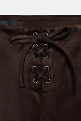 Pantalón de tiro bajo confeccionado en gamuza marrón, con diseño flare y detalle de lazada frontal. Presenta costuras marcadas y cierre lateral con cremallera.