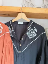 Camisa de color terracota con cuello camisero, cierre frontal de botones y mangas largas abullonadas. Presenta un delicado bordado floral en color blanco en la zona del pecho y los hombros, con un diseño de corte holgado.