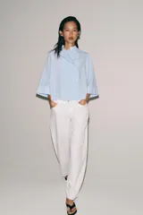 Camisa Zara Woman Collection de algodón con estampado de rayas verticales blancas y celestes. Presenta cuello perkins, mangas tres cuartos y un diseño asimétrico con cierre frontal cruzado mediante botones.