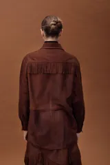 Camisa de cuero ovino color marrón, con diseño oversize, cuello camisero, cierre frontal con broches y detalle de flecos en el pecho. Presenta mangas largas con puños.