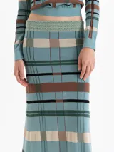 Pollera larga de punto con diseño a cuadros en tonos verde, marrón, negro y beige. Presenta un corte ajustado en la cadera que se ensancha hacia el ruedo y una cintura con detalle de hilo metálico.