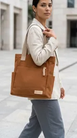 Cartera tipo shopper de lona color marrón, con bolsillos externos laterales, un bolsillo frontal abierto con detalle de aplique y tiras de hombro anchas en tono beige.