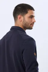 Buzo de felpa color verde oscuro, con cuello tipo polo, cierre frontal parcial con botones y bolsillos laterales. Presenta puños y ruedo acanalados.