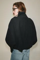 Cazadora bomber negra de gamuza, de cuello subido y manga larga con trabillas en los hombros. Cuenta con bolsillos de plastrón con solapa, cierre frontal con cremallera oculta y botones metálicos a presión, y elástico en el ruedo.