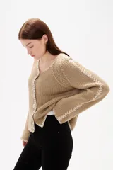 Cardigan de punto color natural, con cuello en V, cierre frontal con botones y diseño holgado. Presenta detalles de costuras decorativas bordadas a lo largo de las mangas y en los bordes.