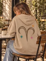 Canguro de felpa color crudo, con capucha, corte amplio, bolsillos delanteros y aberturas laterales en el dobladillo. Presenta un estampado en la espalda con diseño de arco y flecha rodeado de estrellas, y detalles gráficos en las mangas.