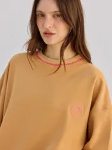Buzo oversize de color tostado con cuello redondo, puños ajustados y detalles bordados en color fucsia en el cuello y el pecho.