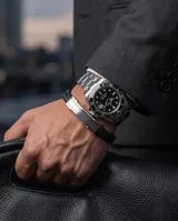 Pulsera masculina de diseño moderno, compuesta por una banda central de acero inoxidable pulido y laterales de cuero trenzado negro. Cuenta con un cierre de seguridad integrado.