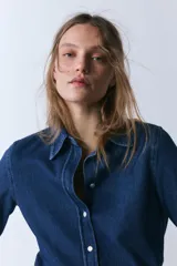 Camisa de denim de corte clásico con cuello solapa, manga larga con puños anchos y cierre frontal con botones. Presenta un diseño de bajo asimétrico.