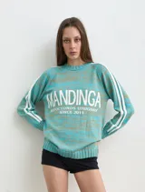 Buzo de punto melange en tonos turquesa y marrón, con cuello redondo, mangas largas ranglán con dos franjas blancas laterales y estampa frontal con la inscripción "MANDINGA AUTOCTONOS URUGUAY SINCE 2011".