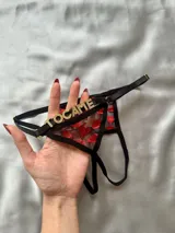 Tanga negra de diseño abierto con tiras regulables, detalles metálicos dorados, aplique de letras en strass con la frase "TOCA ME" en la parte trasera y tejido transparente bordado con corazones rojos.