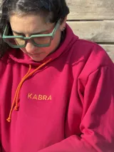 Canguro fucsia de tela deportiva con capucha, cordones ajustables en color naranja y logo bordado en el pecho.